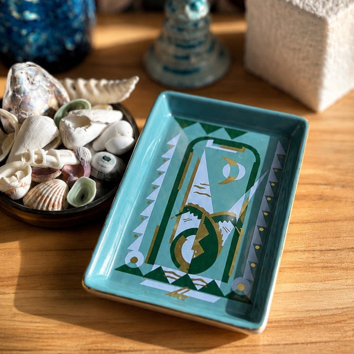 A rectangular turquoise ceramic tray with a geometric green and gold design, styled on a wooden table next to a bowl of seashells and a blue vase.