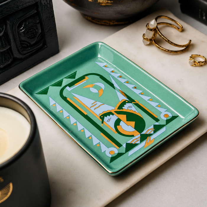 A mint green rectangular tray with a geometric, abstract art print in green, white, and gold, styled on a marble surface with gold rings and a candle nearby.