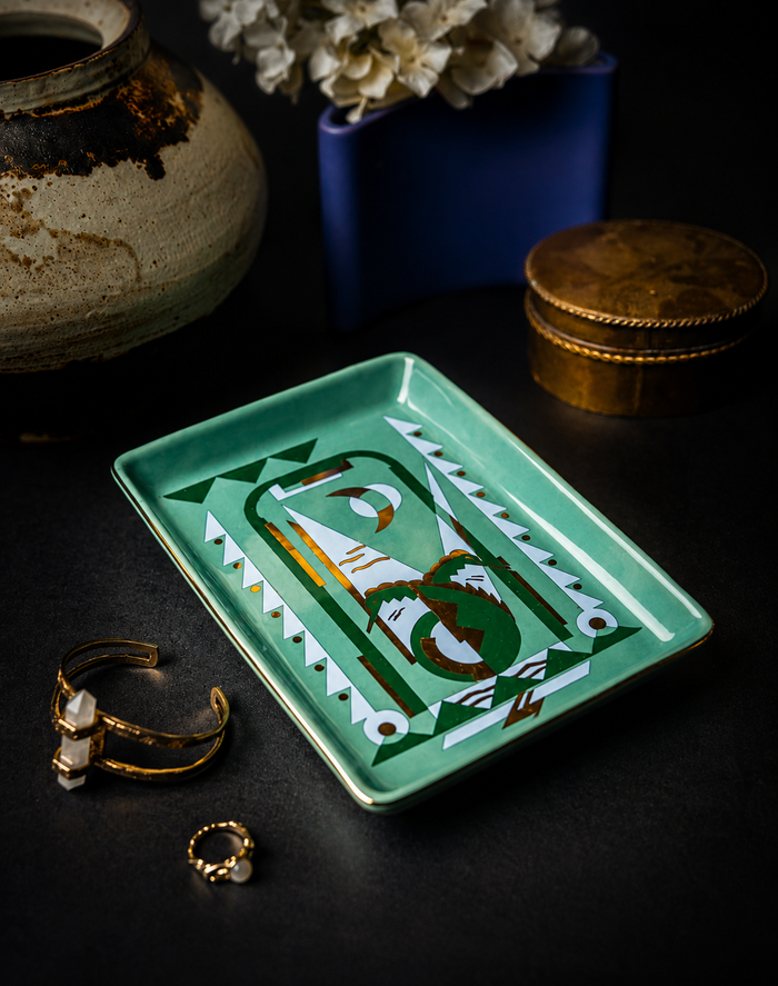 A rectangular mint green ceramic jewelry tray with a hand-painted green, white, and gold geometric design, styled on a dark surface with gold rings and decorative ceramic vases.