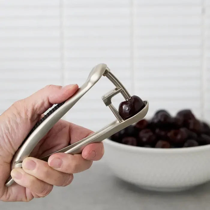 Hand using a cherry pitter to remove pits from cherries with a bowl of cherries in the background.