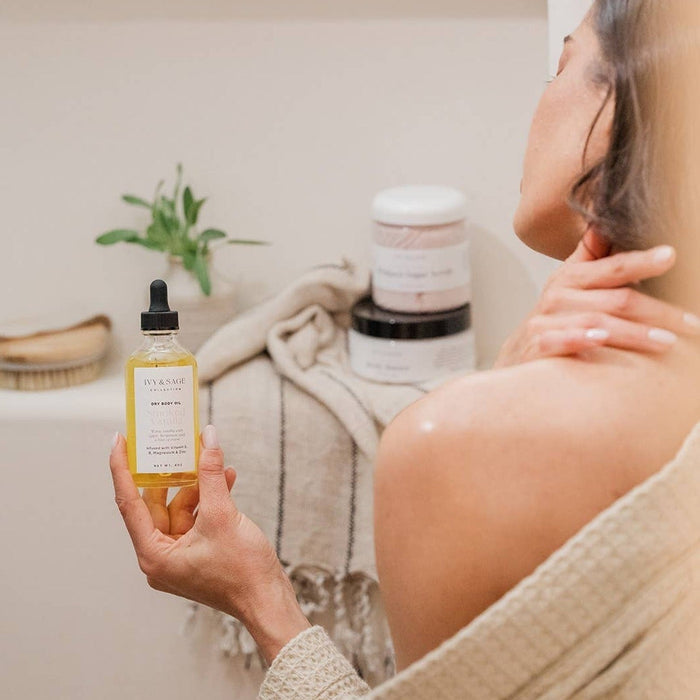 Woman holding Ivy & Sage dry body oil bottle in bathroom setting with skincare products