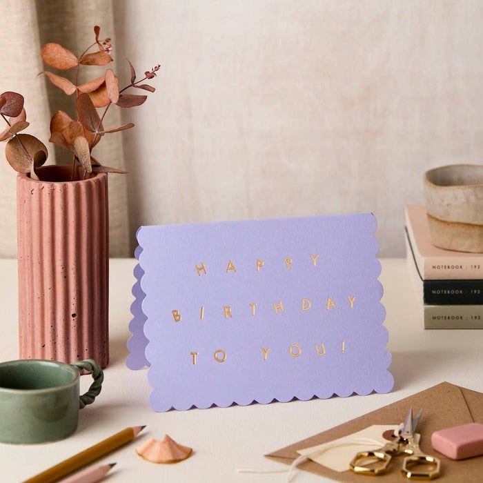 Lavender scallop birthday card displayed on desk with dried flowers, notebooks, and stationery