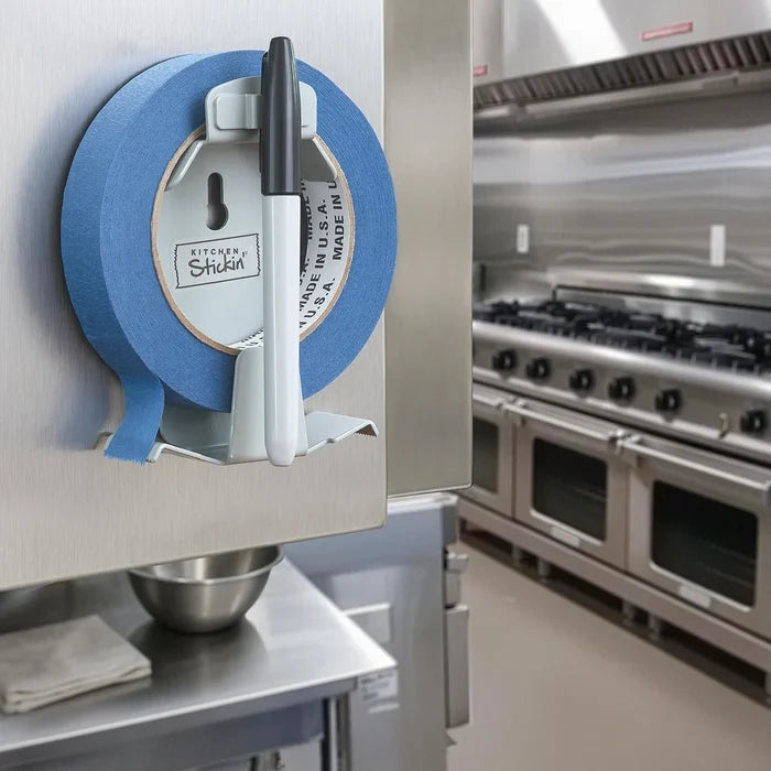 A kitchen marker with a black cap and white barrel is mounted on a kitchen sticken tape dispenser. The background shows a blurred commercial kitchen with stainless steel ovens and a range hood.
