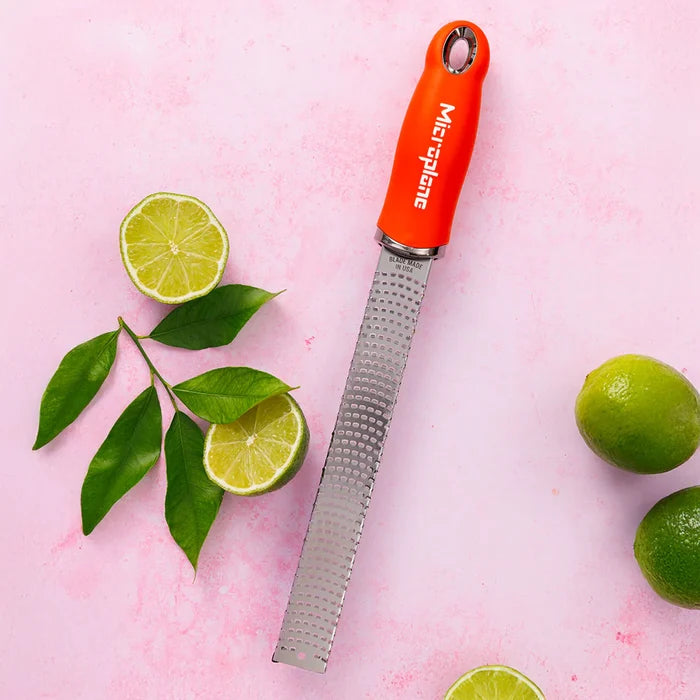 Microplane zester with orange handle and lime halves on a pink background