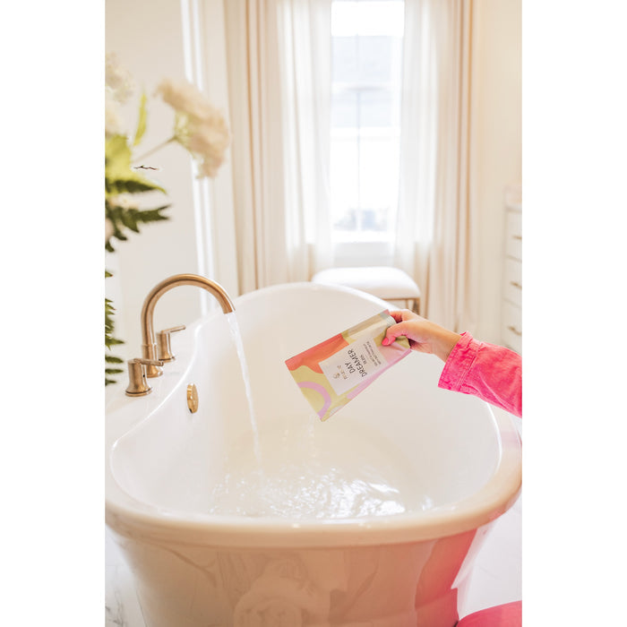 Hand holding Musee Daydreamer Soak bath product over luxurious white soaking tub with running water