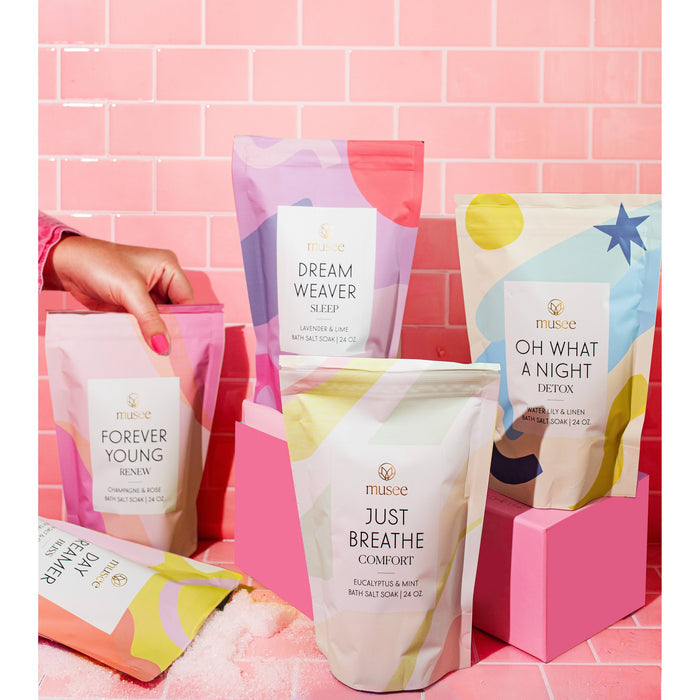 Musee bath salt soaks displayed in colorful packaging with hand reaching toward product on pink brick background