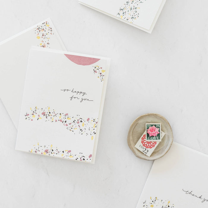 So Happy For You greeting card with terrazzo design, envelope, and love stamps on white surface